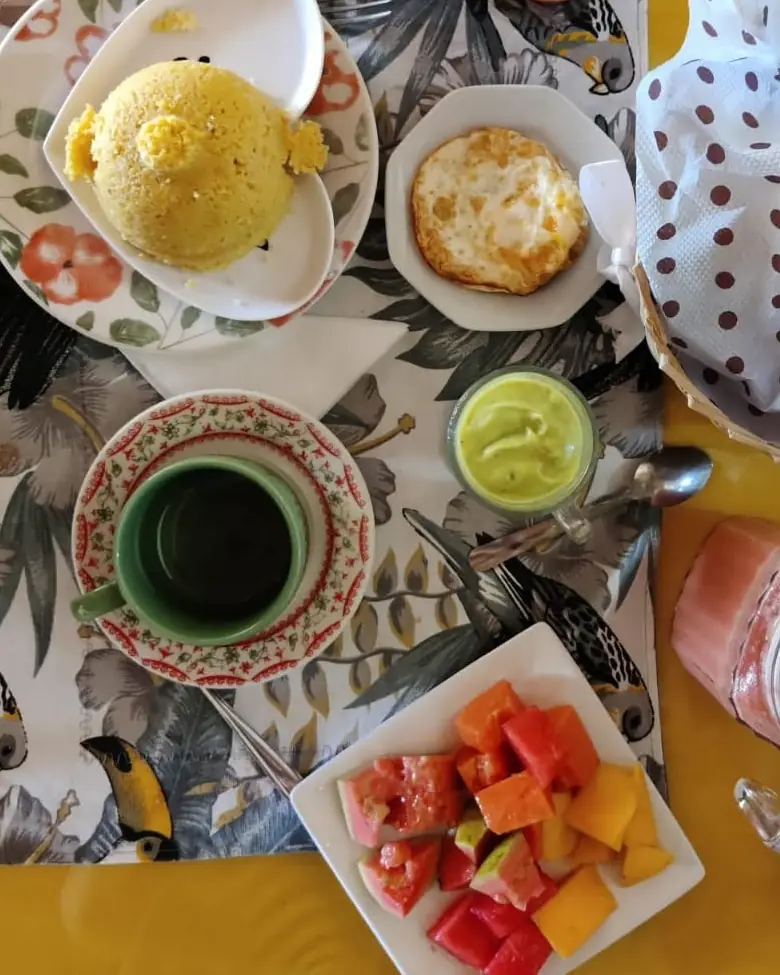 Café da manhã na mesa amarela, visto de cima: cuscuz, ovo, creme de abacate, café, suco e frutas