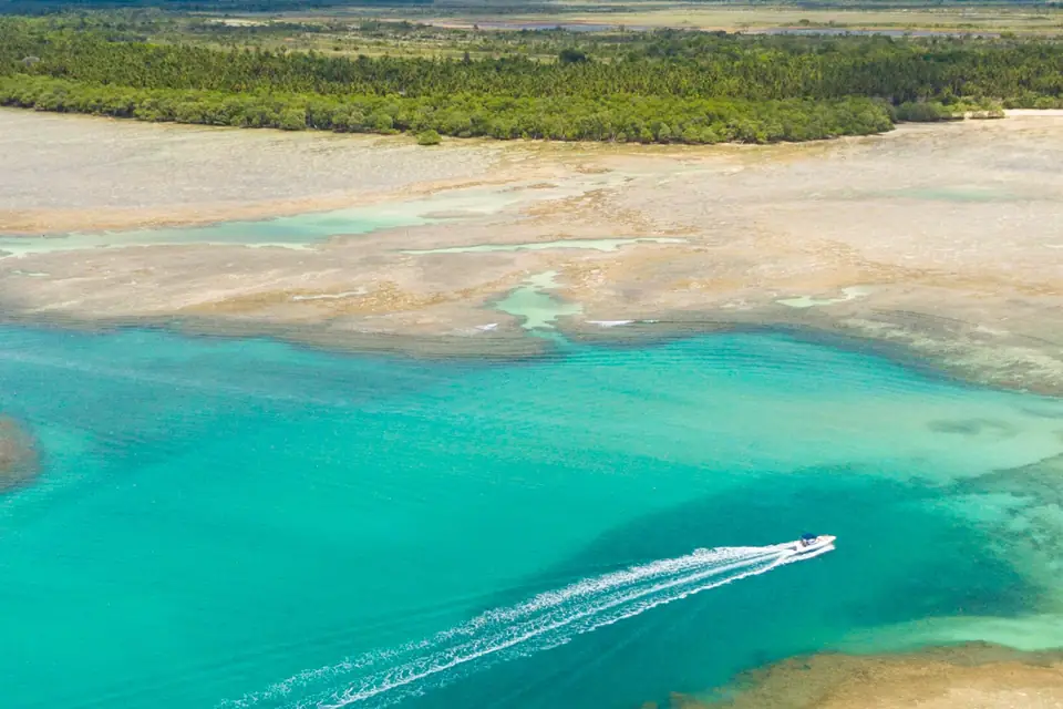 Moreré vista do alto: canal azul-turquesa entre bancos de areia e recifes, uma lancha passando e a mata da ilha ao fundo