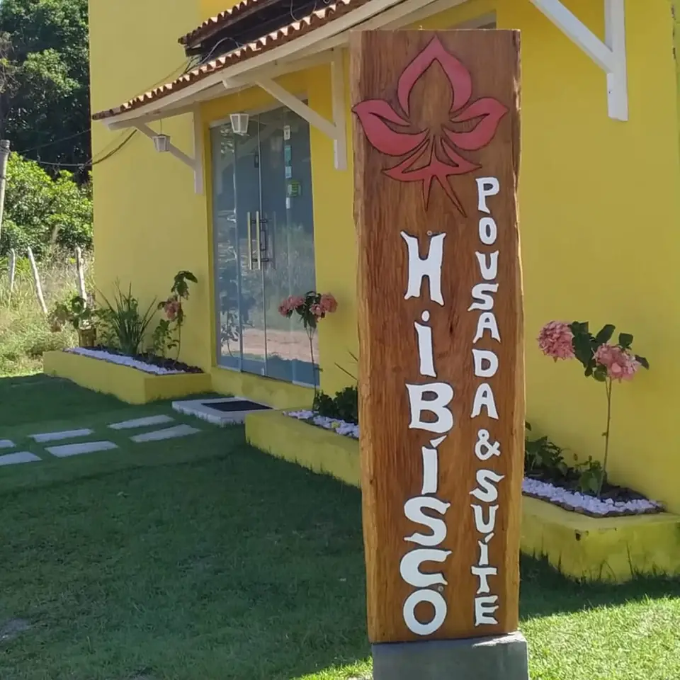 Entrada da pousada: parede amarela, porta de vidro e a placa de madeira entalhada com o hibisco e "Hibisco Pousada & Suíte"